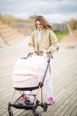 Tam vücut genç anne, dışarıda huzurlu bir gezinti yapar, tahtadan bir kaldırım boyunca bebek arabasıyla gezer.