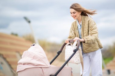 Bej ceketli gülümseyen anne ve beyaz pantolon pembe bebek arabasıyla dışarı çıkıyor.