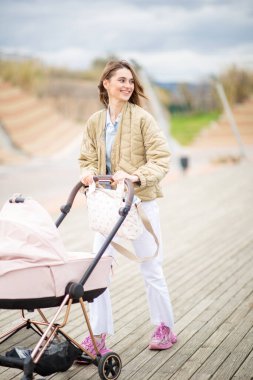 Tüm vücuduyla gülümseyen genç kadın tahta bir tahta kaldırımda yürümekten zevk alıyor, bebek arabasını itiyor.