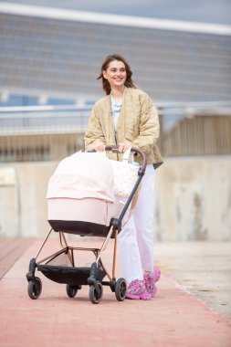 Ceketli genç anne dışarıda yürürken gülümsüyor, zarif pembe bir bebek arabası itiyor..