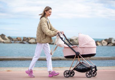 Tam vücut genç anne, bir deniz kenarında huzurlu bir şekilde yürüyor, pembe bir bebek arabasını gülümseyerek itiyor.