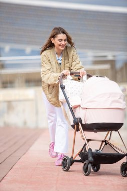 Tüm vücudu neşeli genç anne şehir gezintisinde gezmekten zevk alıyor. Pastel pembe bir bebek arabasını itiyor..