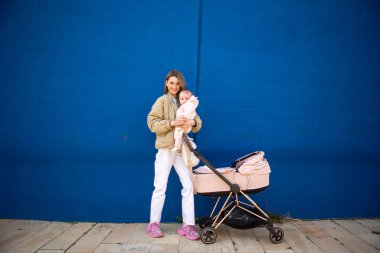 Bebek arabasının yanında bebeğini tutan genç anne mavi duvarın önünde duruyor.
