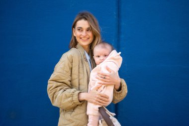 Gülümseyen bir annenin bebeğini pembe elbiseler içinde canlı mavi bir duvara sarışının yakın çekimi.