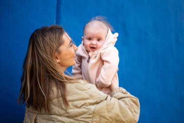 Anne mutlu bebeğini kaldırıyor ve gülümsüyor açık pembe bir elbiseyle, mavi arka planda..