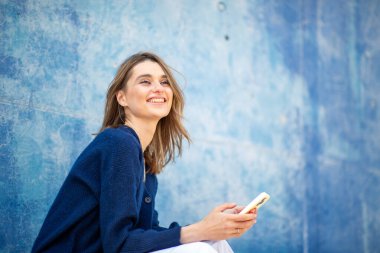 Elinde telefonla gülümseyen genç bir kadın, parlak mavi bir duvarın karşısında oturuyor.