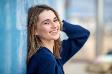 Süveter giymiş gülümseyen bir kadın, bir kolu havada mavi bir duvara yaslanmış.