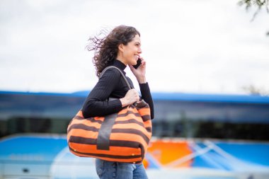 Gülümseyen kadın çantasıyla yürüyor ve arka planda otobüslerle telefonla konuşuyor.