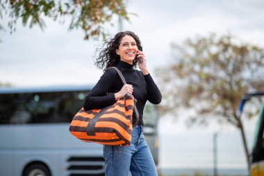 Neşeli bir kadın çantasıyla dışarıda yürüyor, telefonla konuşuyor, park halindeki otobüslerin önünde.