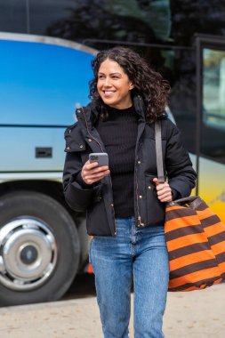Renkli otobüsün yanında yürüyen bir kadın, elinde telefonuyla gülümsüyor.