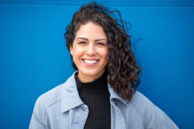 Mavi duvarın önünde duran kıvırcık saçlı neşeli bir kadının portresi.