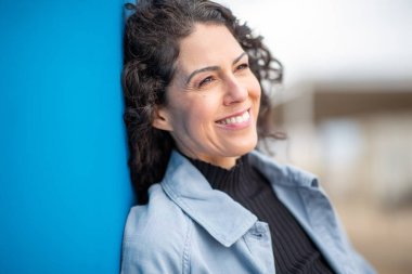 Mavi duvara yaslanmış kıvırcık saçlı, kendine güvenen bir kadın.