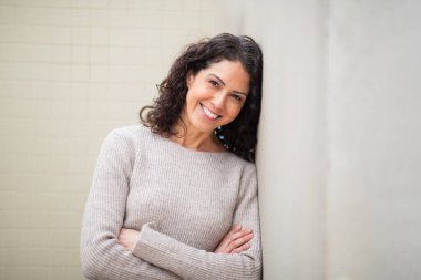 Kollarını kavuşturarak duvara yaslanmış gülümseyen bir kadın portresi.