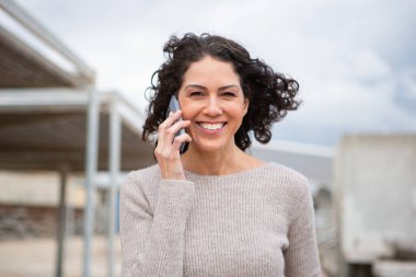 Ön manzara portresi. Kıvırcık saçlı, gülümseyen kadın. Açık havada cep telefonuyla konuşuyor.