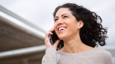 Dışarıda akıllı telefonuyla konuşurken gülümseyen bir kadın. Kıvırcık saçları rüzgarda savruluyor. Yukarı bakıyor.
