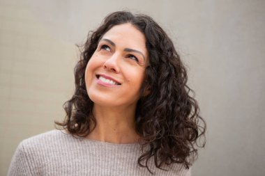 Orta yaşlı, kendinden emin bir kadın. Yukarı bakarken hafifçe gülümsüyor. Tarafsız bir duvarın önünde dikiliyor.