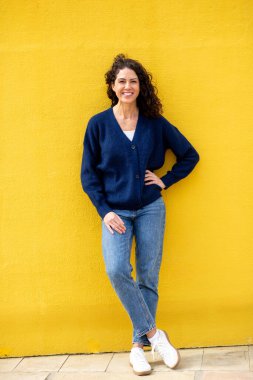 Parlak sarı bir duvara yaslanmış gülümseyen bir kadının tüm vücut portresi.