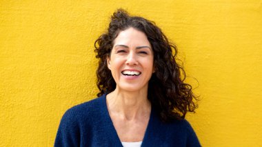 Canlı sarı bir duvarın önünde duran orta yaşlı bir kadının portresi.