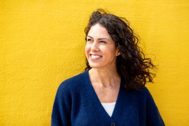 Gülümseyen bir kadının yakın plan fotoğrafı, canlı sarı bir duvara bakıyor.