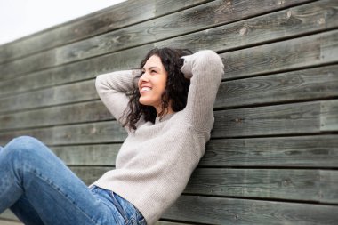 Gülen kadın elleri başının arkasında, kırsal ahşap bir duvara yaslanıyor.