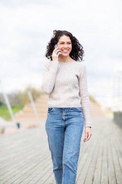 Neşeli kadın ahşap bir kaldırımda yürürken akıllı telefonuyla konuşuyor.