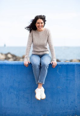 Kadın neşeyle deniz kenarındaki parlak mavi beton çıkıntıda oturuyor.