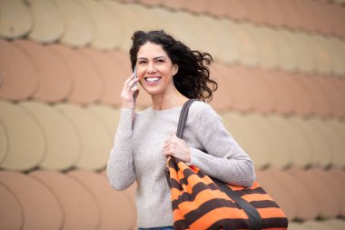 Çantalı gülümseyen kadın dışarıda cep telefonuyla konuşuyor.