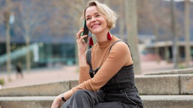 Kot pantolon giymiş gülümseyen kadın dışarıda merdivenlerde otururken cep telefonuyla sohbet ediyor.