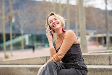 Kot pantolon giymiş orta yaşlı bir kadın dışarıda merdivenlerde otururken cep telefonuyla sohbet ediyor.