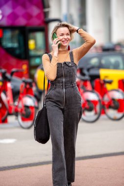 Gülümseyen bir kadın telefonda konuşurken şehirde yürüyor.