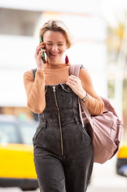 Sarışın, gülümseyen bir kadın. Şehirde yürürken telefonla konuşuyor.