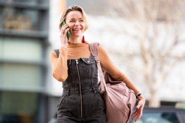 Sarışın, gülümseyen kadın şehirde yürürken akıllı telefonuyla konuşuyor.