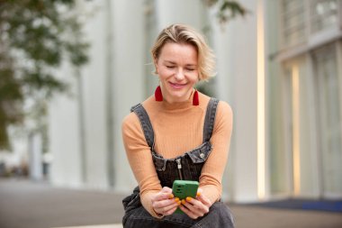 Neşeli kadın dışarıda oturmuş, gülümseyerek akıllı telefonuna bakıyor.