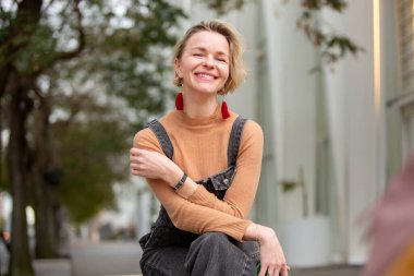 Kadın dışarıda oturuyor, kameraya sıcak bir şekilde gülümsüyor. Rahatlamış vücut dili ve kendine güveni insana neşe ve rahatlık veriyor.