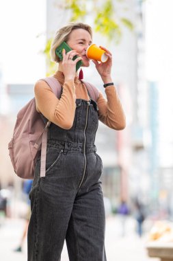 Sarışın bir kadının elinde içki ve cep telefonuyla şehirde yürümesi.