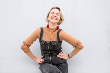 Beyaz bir duvara karşı poz verirken gülümseyen sarışın kadın.