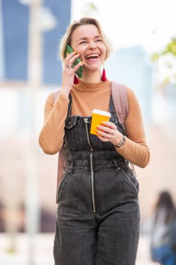 Sarışın kadın gidiyor, cep telefonu ve bir içkiyle yürüyor.