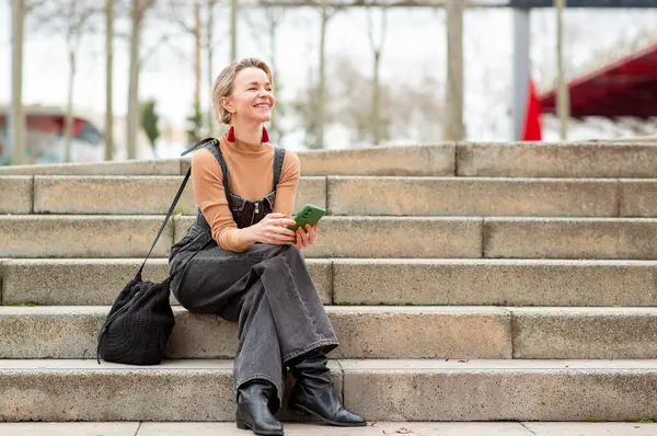 Neşeli kadın beton bir merdivende rahatça oturur, elinde akıllı telefon tutarak.
