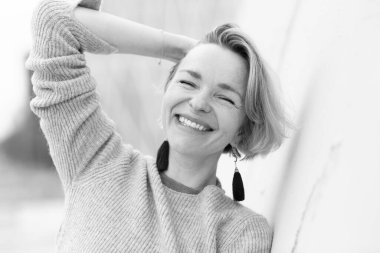 Mutlu sarışın kadın gözleri kapalı gülüyor, beyaz bir duvara yaslanıyor.