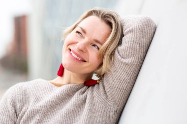 Açık havada duvara yaslanmış gülümseyen sarışın kadın bej kazak ve kırmızı püsküller takıyor.