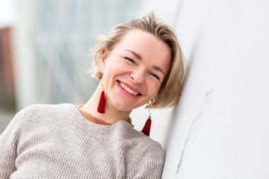 Gülümseyen sarışın kadın şakacı bir şekilde beyaz bir duvara yaslanıyor.