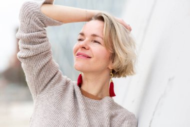 Gözleri kapalı gülümseyen kendine güvenen sarışın kadın, beyaz bir duvara yaslanıyor.