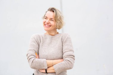 Süveter giyen gülümseyen sarışın kadın kollarını beyaz arka planda kavuşturuyor.