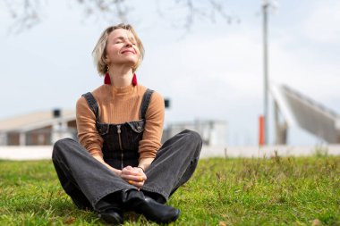 Kadın çimenlerin üzerinde bağdaş kurmuş oturuyor, gözleri kapalı ve gün ışığından zevk alırken huzur içinde gülümsüyor.