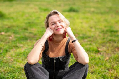 Sarışın kadın güneşli bir açık havada çimlerin üzerinde oturuyor, çenesi elleri üzerinde gülümsüyor.