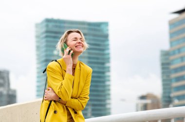 Sarı ceketli neşeli bir kadın şehirde cep telefonuyla konuşurken gülüyor. Kadın, kadın, insan, bir yetişkin, sarışın, kısa saçlı, gülümseyen, mutlu, neşeli, pozitif, neşeli, kendine güvenen, çekici, modaya uygun, modern