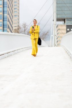 Sarı takım elbiseli bir kadın modern bir köprüde yürüyor ve elinde cep telefonu var.