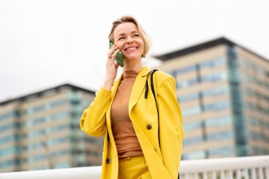 Sarı takım elbiseli, gülümseyen kadın cep telefonuyla konuşuyor..