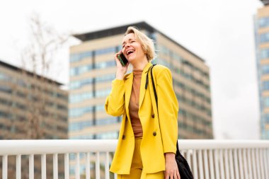 Sarı takım elbiseli bir kadın cep telefonuyla konuşurken köprüde yürüyor..