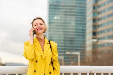 Sarı ceketli gülümseyen kadın arka planda yüksek şehir binaları olan açık havada cep telefonuyla konuşuyor..